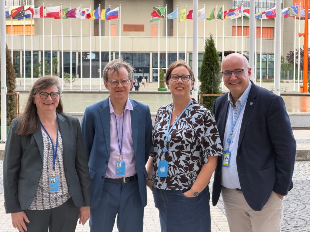 Four professionals wearing lanyards stand shoulder-to-shoulder, smiling outdoors on a paved plaza with a line of international flags in the background.