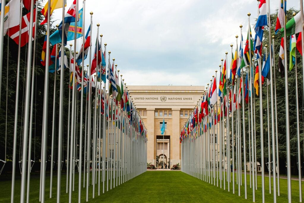 Two long parallel rows of national flags on tall silver poles flank a green lawn leading toward the United Nations building in Geneva.