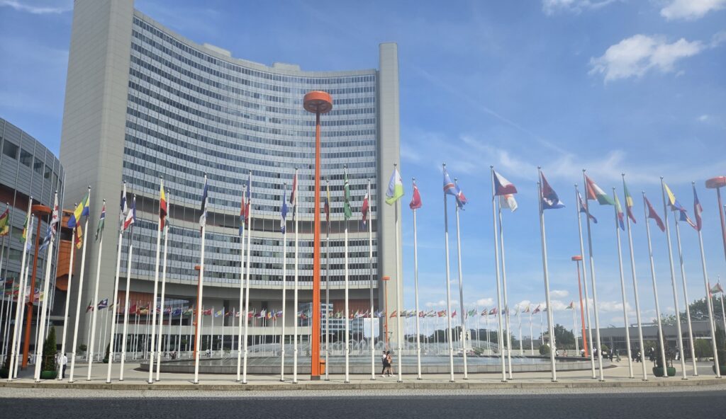 A wide shot of a curved, modern high-rise building with a large fountain plaza in front, lined with numerous national flags on tall poles under a bright blue sky.