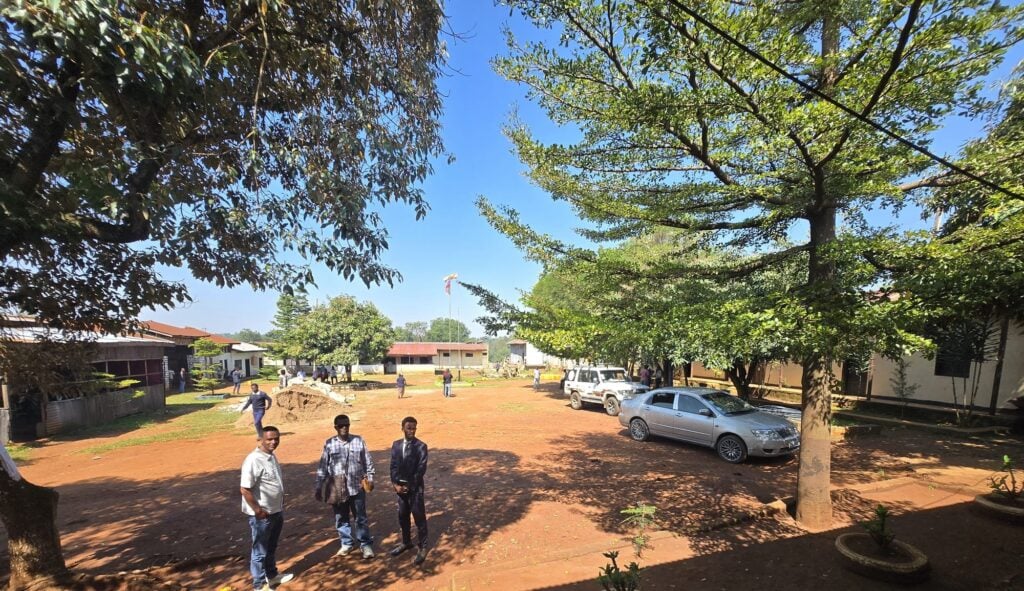A sunny dirt yard with trees, parked cars and low buildings outside a prison in Ethiopia, with a few men standing in the foreground